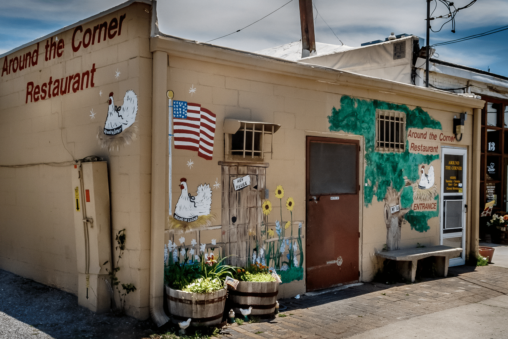 A small restaurant called "Around the Corner Restaurant," often listed among the 10 best restaurants in Edmond OK, features a mural with chickens, an American flag, sunflowers, and the word "Entrance" on the right side. The tan building is adorned with potted plants, creating a cozy atmosphere.