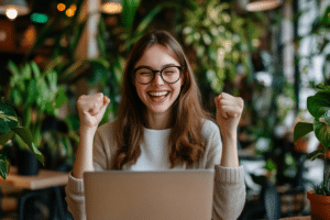 Do you really need a real estate agent? A woman with glasses and a wide smile raises her fists in excitement while sitting in front of a laptop in a plant-filled room.