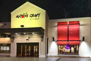 Exterior of Anton's Craft Kitchen at night, with a prominent sign and red window coverings, and an illuminated "open" sign visible inside.