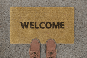 A pair of brown shoes standing in front of a tan doormat with the word "WELCOME" on a gray, speckled concrete surface.