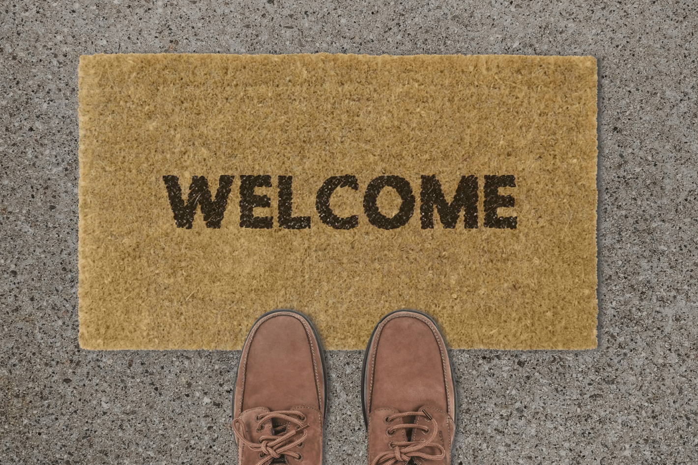 Copy of BAMx in a Box Blog - 06-20-25 A pair of brown shoes standing in front of a tan doormat with the word "WELCOME" on a gray, speckled concrete surface.