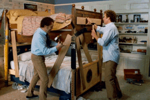 Two men in a messy bedroom attempt to build a bunk bed using wooden planks and tools, with personal items and sports memorabilia on shelves in the background.