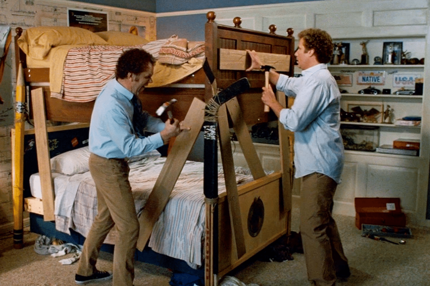 getting-creative-to-afford-a-home Two men in a messy bedroom attempt to build a bunk bed using wooden planks and tools, with personal items and sports memorabilia on shelves in the background.