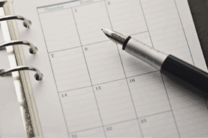 A fountain pen rests on an open ring-bound planner, displaying a blank weekly calendar page.