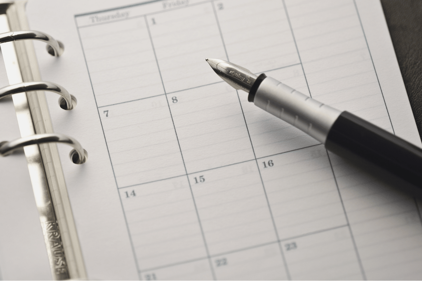 A fountain pen rests on an open ring-bound planner, displaying a blank weekly calendar page.