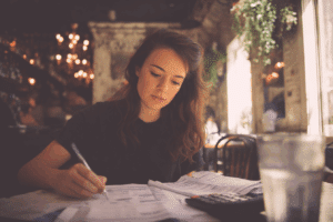 A woman sits at a table in a cafe, focused on paperwork with a pen in hand, a calculator, and a glass of water nearby.