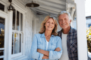 A couple stand side by side and smile on the porch of a house on a sunny day as they consider getting their home ready to sell.