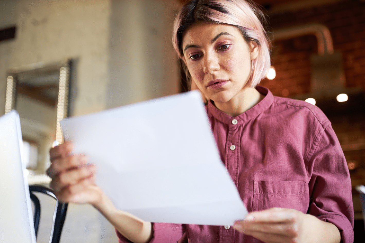 A woman with pink hair wearing a mauve shirt looks intently at a sheet of paper she is holding indoors.