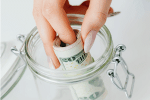A hand with manicured nails places a rolled-up U.S. twenty-dollar bill into a clear glass jar with a hinged lid.