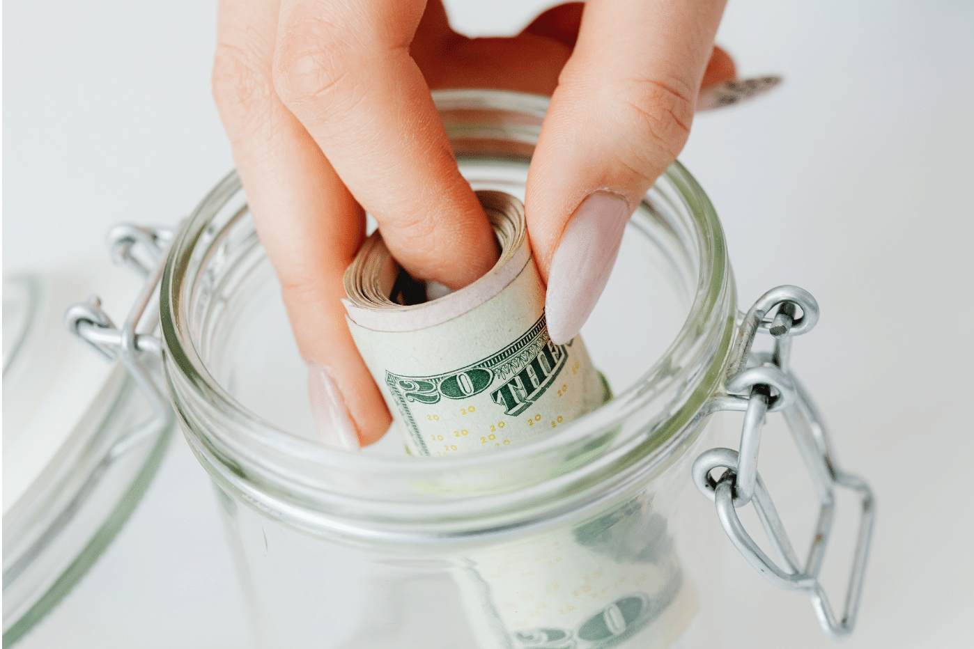 A hand with manicured nails places a rolled-up U.S. twenty-dollar bill into a clear glass jar with a hinged lid.
