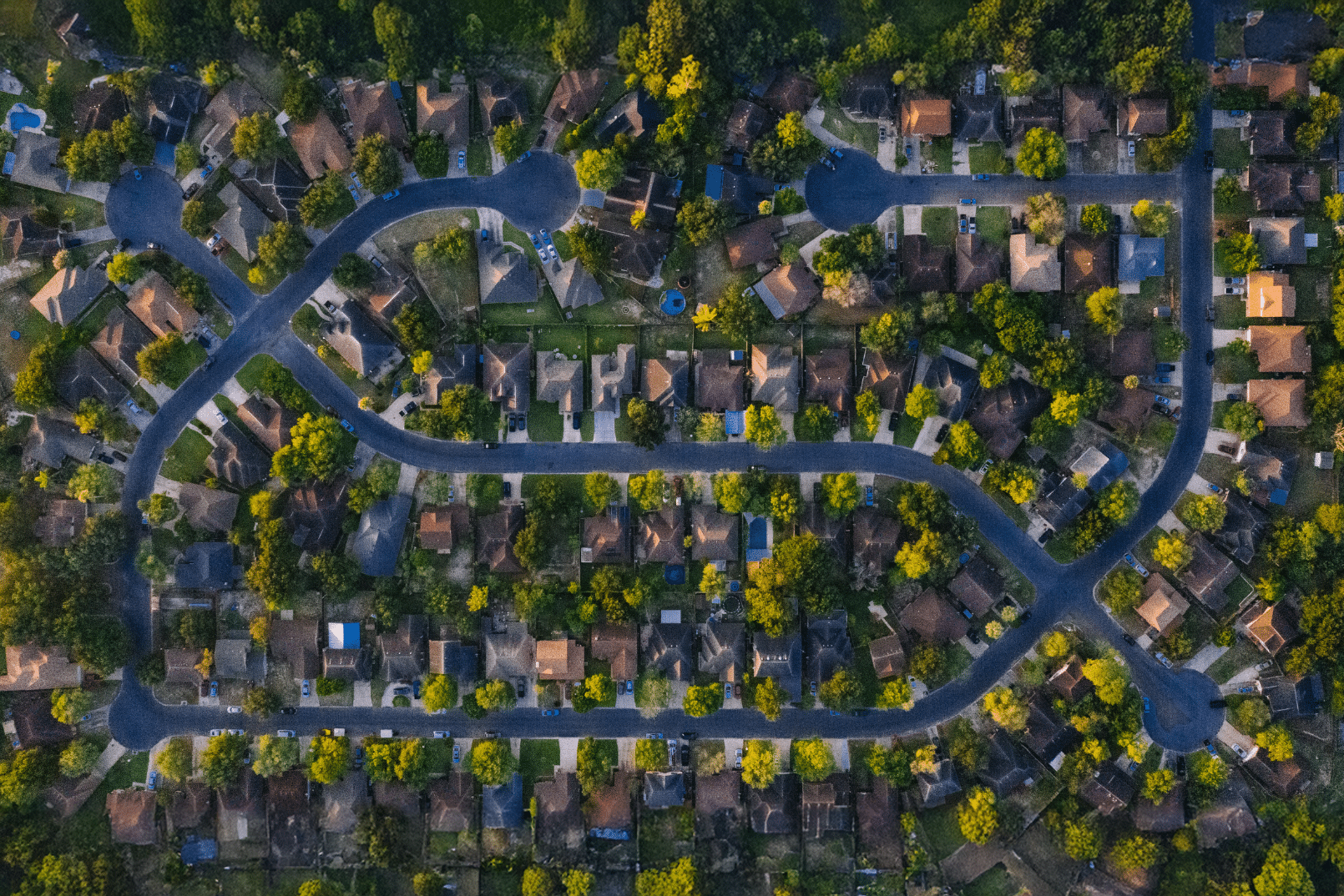 Aerial view of a suburban neighborhood showing curved streets, houses with driveways, and scattered green trees.