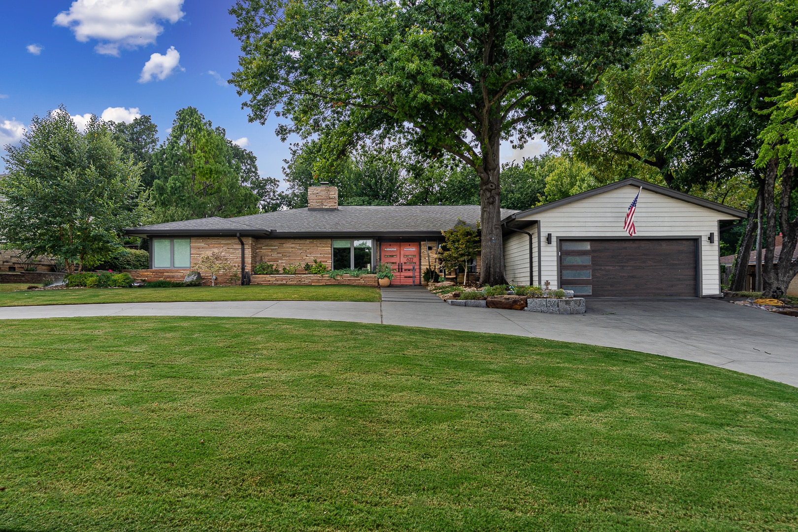 Single-story house with stone and siding exterior, attached garage, curved driveway, well-manicured lawn, mature trees, and an American flag—this inviting property at 622 Timber Lane is a prime example of Clegern Edmond Oklahoma homes for sale. Enjoy outdoor entertaining beneath the steel pavilion outdoor kitchen nestled amid lush greenery.
