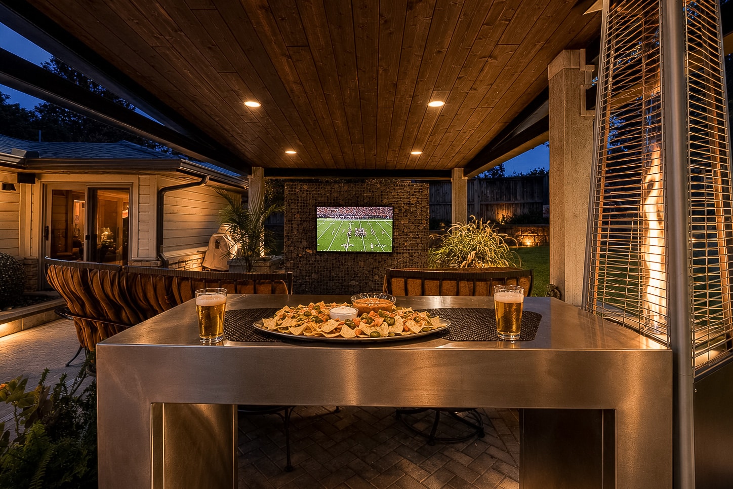 Beneath the wooden roof at dusk, this inviting outdoor patio at 622 Timber Lane Edmond OK features comfortable seating, a TV showing a football game, and a table set with food and two beers in the foreground. Experience relaxed evenings and sophisticated entertaining in this beautifully rebuilt mid-century modern home—one of the standout Clegern Edmond Oklahoma homes for sale.
