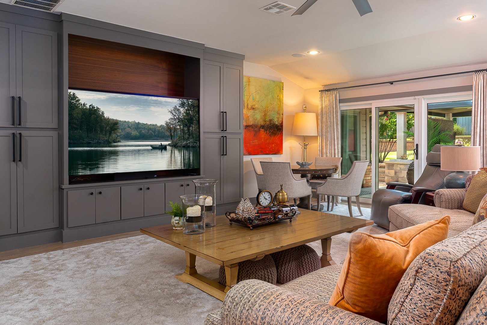 Modern living room with gray built-in cabinets, large TV displaying a lake scene, wooden coffee table, sofa with orange pillows, and dining area near glass doors to a patio—showcasing the inviting style often found in Clegern Edmond Oklahoma homes for sale.