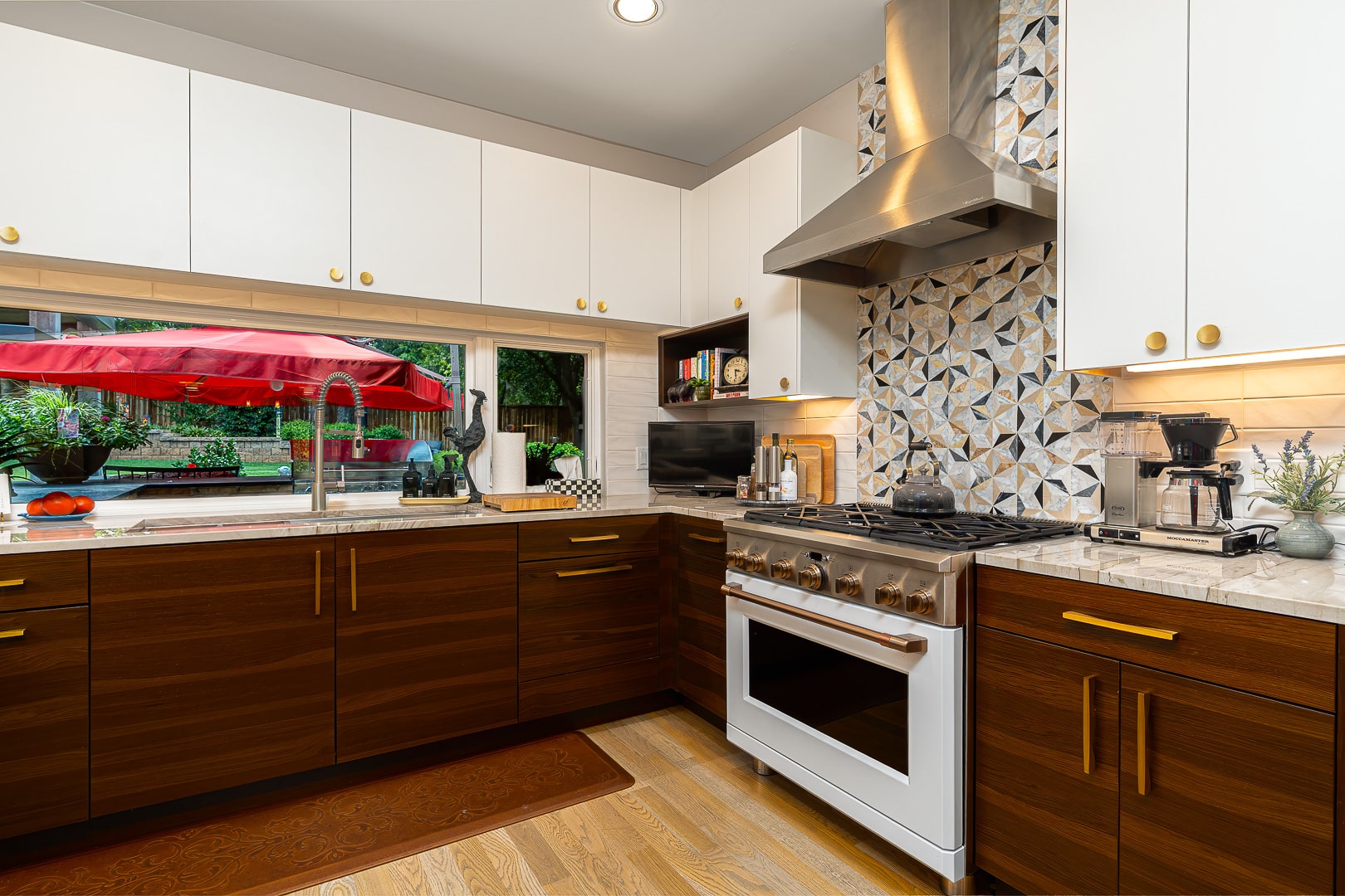 Modern kitchen with white and wood cabinets, stainless steel appliances, geometric tile backsplash, and a window overlooking a patio with a red umbrella—perfectly capturing the airy vibe of a rebuilt mid-century modern home. This inviting space is ideal for those seeking Clegern Edmond Oklahoma homes for sale.