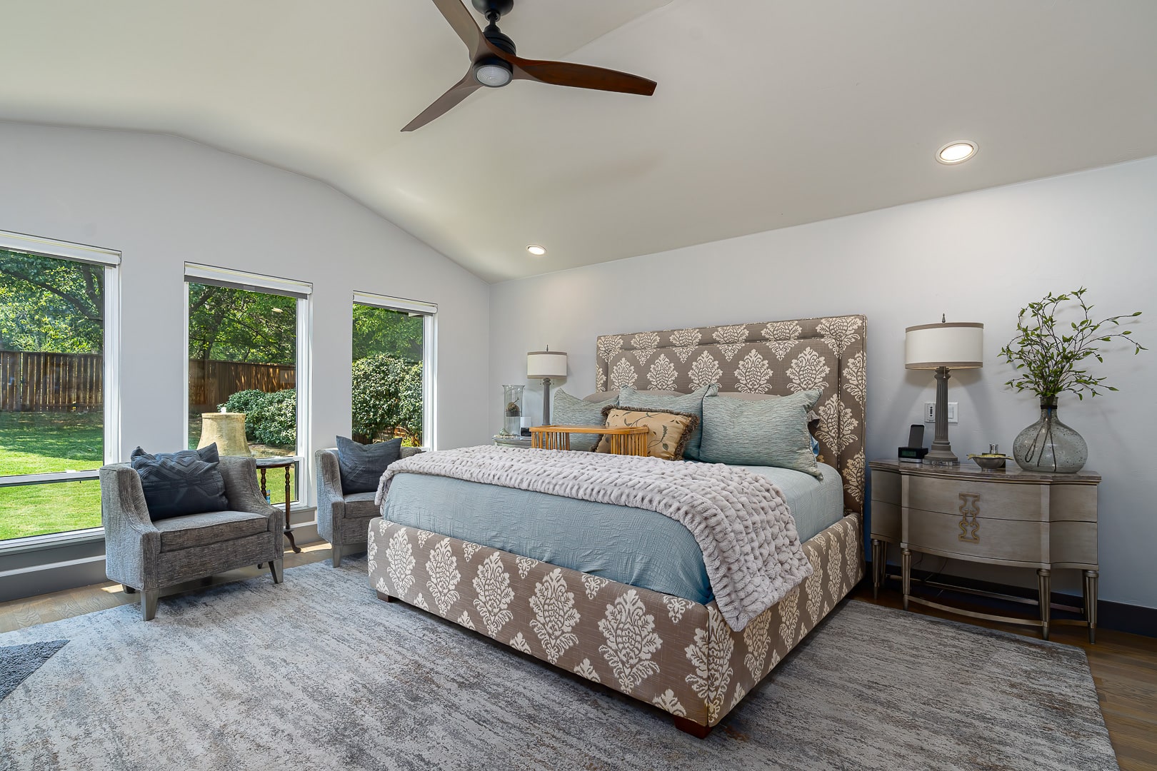 A spacious bedroom at 622 Timber Lane Edmond OK features a patterned bed, light blue bedding, two gray chairs, nightstands with lamps, and large windows overlooking a green backyard—one of the inviting spaces found in Clegern Edmond Oklahoma homes for sale.