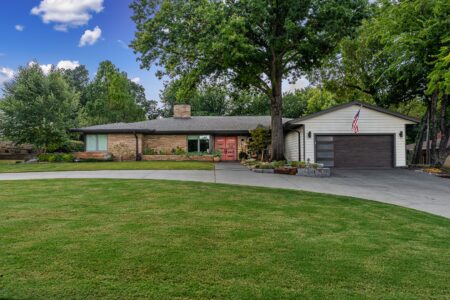 Single-story house with stone and siding exterior, attached garage, curved driveway, well-manicured lawn, mature trees, and an American flag—this inviting property at 622 Timber Lane is a prime example of Clegern Edmond Oklahoma homes for sale. Enjoy outdoor entertaining beneath the steel pavilion outdoor kitchen nestled amid lush greenery.