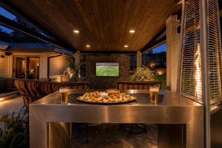 Beneath the wooden roof at dusk, this inviting outdoor patio at 622 Timber Lane Edmond OK features comfortable seating, a TV showing a football game, and a table set with food and two beers in the foreground. Experience relaxed evenings and sophisticated entertaining in this beautifully rebuilt mid-century modern home—one of the standout Clegern Edmond Oklahoma homes for sale.