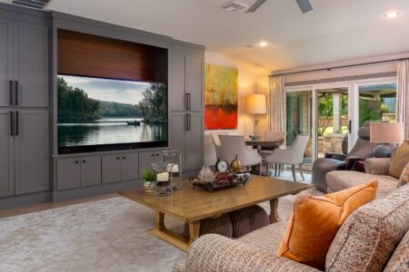 Modern living room with gray built-in cabinets, large TV displaying a lake scene, wooden coffee table, sofa with orange pillows, and dining area near glass doors to a patio—showcasing the inviting style often found in Clegern Edmond Oklahoma homes for sale.