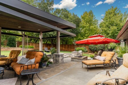 Modern backyard patio with cushioned seating, a stone fire pit, red umbrellas, and trees in the background under a blue sky—perfect for enjoying the outdoors at 622 Timber Lane Edmond OK, a beautifully rebuilt mid-century modern home.