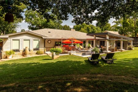 Single-story house with a large backyard, shaded patio, outdoor seating, and two lounge chairs on a grassy lawn under trees. Located at 622 Timber Lane Edmond OK in the desirable Clegern neighborhood, this home also features a stylish steel pavilion outdoor kitchen—perfect for entertaining or relaxing outdoors.