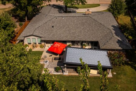 Aerial view of 622 Timber Lane Edmond OK, a beautifully rebuilt mid-century modern home with a gray roof, a red patio umbrella, and surrounding trees in a residential neighborhood—featuring a stylish steel pavilion outdoor kitchen perfect for entertaining.
