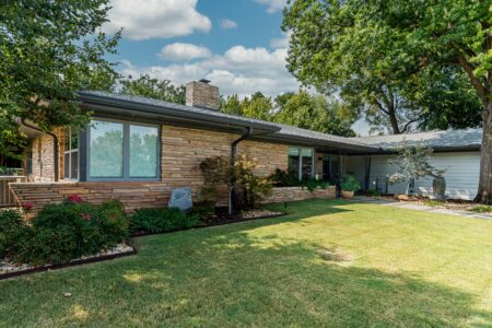 Single-story rebuilt mid-century modern home with stone facade, large windows, and manicured lawn, surrounded by trees and shrubs under a partly cloudy sky.