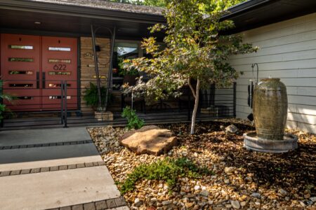 Modern house entry with red double doors, stone steps, a small tree, a rock water fountain, and a landscaped yard with gravel and mulch. This beautifully rebuilt mid-century modern home features a stunning steel pavilion outdoor kitchen—perfect for entertaining—and is among the standout Clegern Edmond Oklahoma homes for sale.