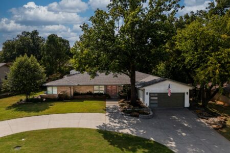 Single-story house with a two-car garage, large driveway, green lawn, and mature trees under a partly cloudy sky. An American flag hangs near the garage door. Located at 622 Timber Lane Edmond OK, this home also features a steel pavilion outdoor kitchen—perfect for entertaining outdoors.