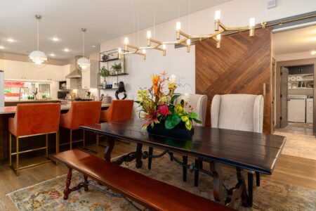 Modern dining room with a dark wood table, beige chairs, floral centerpiece, orange barstools at a kitchen island, and a gold chandelier. A laundry room is visible through an open sliding door. This inviting space is part of 622 Timber Lane Edmond OK, one of the sought-after Clegern Edmond Oklahoma homes for sale.