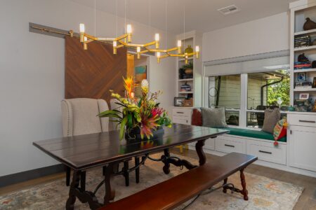A modern dining room in this rebuilt mid-century modern home at 622 Timber Lane Edmond OK, featuring a dark wooden table, bench seating, floral centerpiece, built-in shelves, a large window, and a wooden sliding barn door.