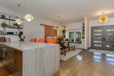 Modern open-concept kitchen and dining area with marble island, orange chairs, pendant lighting, wooden floors, and a dark front door—an inviting highlight at 622 Timber Lane Edmond OK. This residence exemplifies the stylish comfort found in many Clegern Edmond Oklahoma homes for sale.